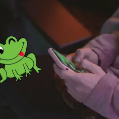 Zielona żaba z wywieszonym językiem obok dłoni trzymających smartfon.